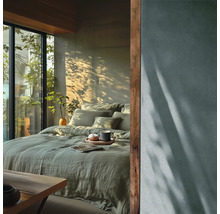 Modernes Schlafzimmer mit grüner Leinenbettwäsche, großem Fenster mit Blick ins Grüne und natürlichem Licht mit Pflanzenschatten an der Wand.