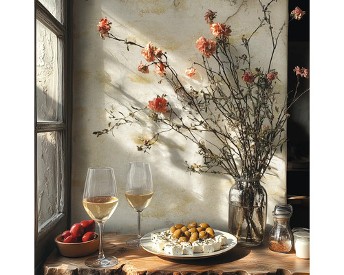 Rustikaler Tisch am Fenster mit Weißwein, Oliven, Fetakäse, Tomaten und einer Vase mit Trockenblumen vor einer texturierten Wand.
