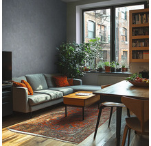 Modernes Wohnzimmer mit grauer Wand in Betonoptik, grauem Sofa, Holztisch, orientalischem Teppich und großen Zimmerpflanzen vor einem Fenster.