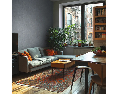 Modernes Wohnzimmer mit grauer Wand in Betonoptik, grauem Sofa, Holztisch, orientalischem Teppich und großen Zimmerpflanzen vor einem Fenster.
