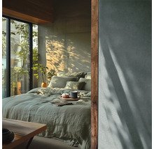 Modernes Schlafzimmer mit grüner Leinenbettwäsche, großer Fensterfront zum Garten und einer Wand in Betonoptik mit Licht- und Schattenspielen.