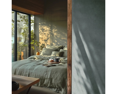 Modernes Schlafzimmer mit grüner Leinenbettwäsche, großer Fensterfront zum Garten und einer Wand in Betonoptik mit Licht- und Schattenspielen.