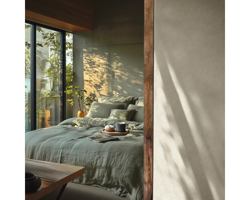 Modernes Schlafzimmer mit Bett in grüner Leinen-Bettwäsche, Wand in Betonoptik mit Baumschatten und großes Fenster mit Pflanzen.