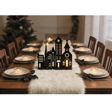 Festlich gedeckter Holztisch mit schwarzer Stadtsilhouette, weißem Fellläufer, Tellern mit Teelichtern und Stechpalmen. Weihnachtsbaum im Hintergrund.