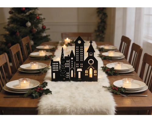 Festlich gedeckter Holztisch mit schwarzer Stadtsilhouette, weißem Fellläufer, Tellern mit Teelichtern und Stechpalmen. Weihnachtsbaum im Hintergrund.