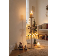 Hoher Metallkerzenständer mit dekorativer Schleife und brennender Kerze in einem weihnachtlich dekorierten Wohnzimmer mit Weihnachtsbaum und Figuren.