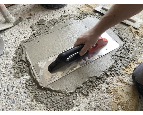 Eine Hand trägt mit einer Zahnkelle Mörtel oder Fliesenkleber auf einen rauen Betonboden auf.