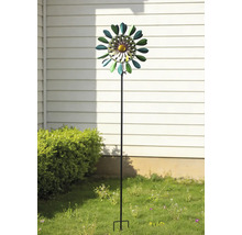 Windrad aus Metall in Blumenform auf einem Gartenstab, platziert in einem Garten vor einer Hauswand und einem Gebüsch.