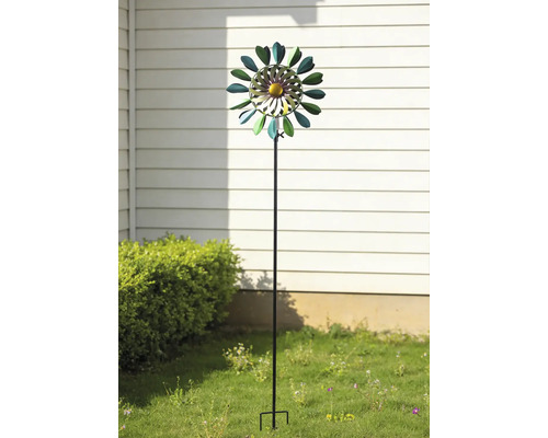 Windrad aus Metall in Blumenform auf einem Gartenstab, platziert in einem Garten vor einer Hauswand und einem Gebüsch.