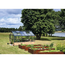 Gewächshaus mit schwarzem Aluminiumrahmen auf einem Ziegelsockel in einem Garten mit Holz-Hochbeeten, Bäumen und einem See im Hintergrund.