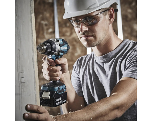 Ein Mann mit weißem Schutzhelm und Schutzbrille verwendet einen Makita 18 Volt Akku-Bohrschrauber, um in ein Holzbrett zu bohren.