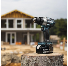 Makita Akku-Bohrschrauber 18 Volt mit 6,0 Amperestunden Akku auf einem Baumstamm vor einer Hausbaustelle.