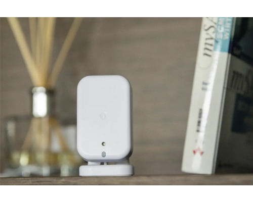 Weißer Smart Home Bewegungsmelder auf einem Regal neben einem Buch und einem Raumduft