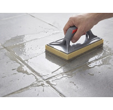 Reinigung von Bodenfliesen mit einem Schwammbrett mit grauem Griff und gelbem Schwammbelag nach dem Verfugen.