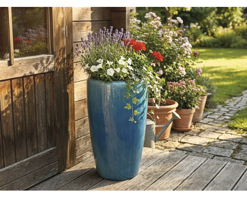Hoher blauer Keramik-Pflanztopf bepflanzt mit Lavendel und Geranien auf einer Holzterrasse neben einem hölzernen Gartenhaus.