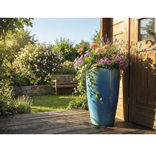 Hoher blauer Keramik-Pflanzkübel mit Sommerblumen und Efeu auf einer Holzterrasse in einem hellen Garten mit Gartenbank und Steinmauer.
