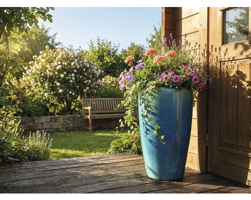 Hoher blauer Keramik-Pflanzkübel mit Sommerblumen und Efeu auf einer Holzterrasse in einem hellen Garten mit Gartenbank und Steinmauer.