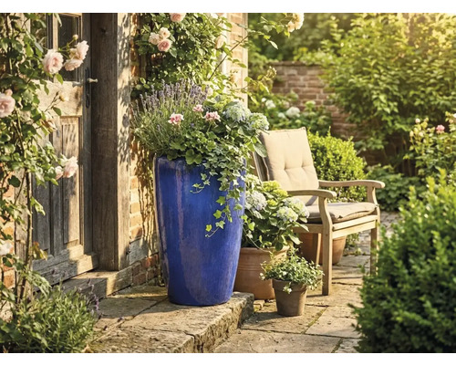 Hoher blauer Keramik-Pflanzkübel auf Steinterrasse neben rustikaler Holztür, Gartenstuhl und Blumen im hellen, gepflegten Garten.