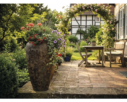 Bepflanztes hohes Pflanzgefäß auf einer gepflasterten Terrasse mit Gartenmöbeln aus Holz, Rosenbogen und Fachwerkhaus im Hintergrund.