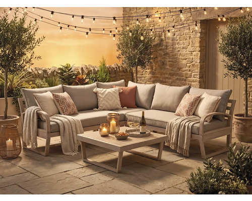 Ecklounge-Set aus Holz mit grauen Polstern, Couchtisch, Kissen, Decken, Lichterkette und Laternen auf einer Steinterrasse vor Steinmauer.