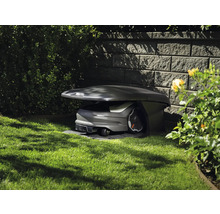 Husqvarna Mähroboter in einer Garagenstation auf einer Rasenfläche neben einer Steinmauer im Garten.