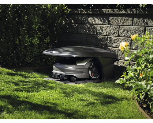 Husqvarna Mähroboter in einer Garagenstation auf einer Rasenfläche neben einer Steinmauer im Garten.