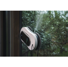 LifenaXX Fensterreinigungsroboter reinigt eine Fensterscheibe mit integrierter Sprühfunktion vor einem Gartenhintergrund.