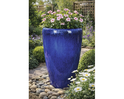 Hoher, blau glasierter Pflanzkübel aus Keramik mit rosa Blüten in einer Gartenumgebung mit Kieselsteinen.