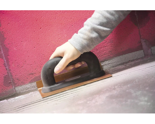 Eine Hand führt ein Reibebrett über eine Bodenfläche entlang einer strukturierten Wand.