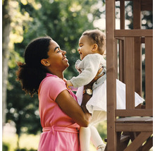 Frau hält ein Kleinkind vor einem Spielturm aus Holz im Garten.