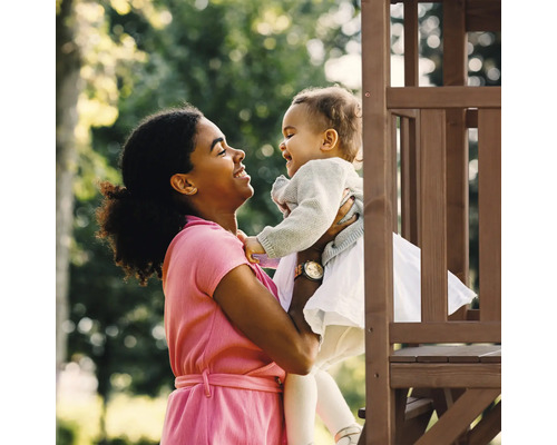 Frau hält ein Kleinkind vor einem Spielturm aus Holz im Garten.