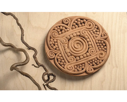 Runde Ton-Dekoration mit Spiralmustern und Relief-Verzierungen auf einer Holzoberfläche neben einem gewundenen Ast.