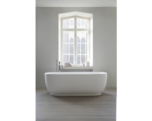 Freistehende weiße Badewanne in modernem hellem Badezimmer vor einem Fenster, bodenstehende Armatur, Holzfußboden.