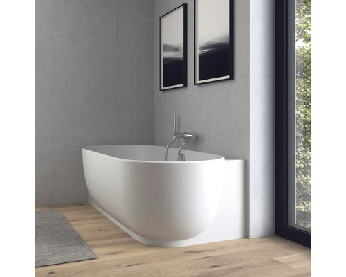 Moderne, ovale Badewanne in Weiß mit Wandarmatur aus Chrom in einem hellen Badezimmer mit Betonoptik-Wänden und Holzparkett.