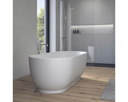Freistehende, ovale Badewanne in einem modernen Badezimmer mit Wänden in Betonoptik, Holzboden und einer Dusche im Hintergrund.