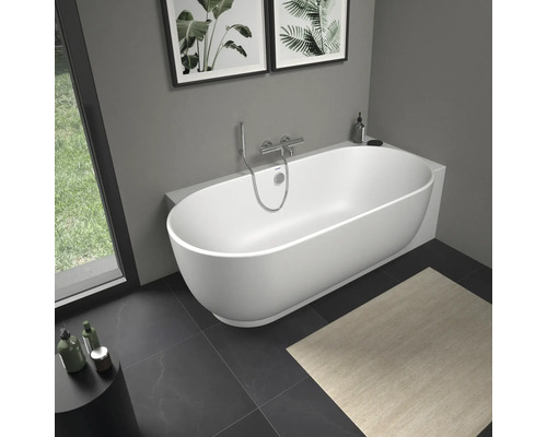 Moderne freistehende weiße Badewanne in ovaler Form in einem hellen Badezimmer mit Wandarmatur aus Chrom und dunklen Bodenfliesen.