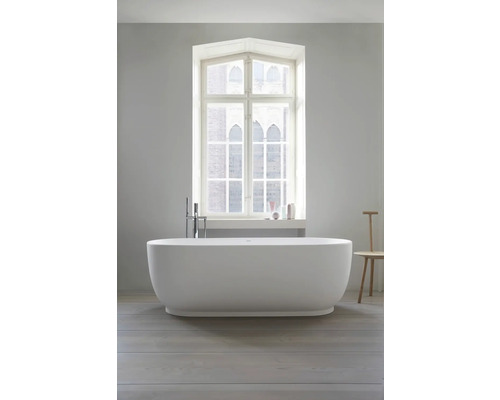 Freistehende ovale Badewanne mit bodenstehender Armatur vor einem großen Fenster in einem hellen, modernen Badezimmer.
