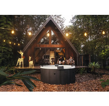 Runder Außenwhirlpool in einem Garten mit modernem A-Frame-Haus, Lichterketten und leuchtenden Rentier-Dekorationen am Abend.