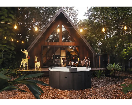 Runder Außenwhirlpool in einem Garten mit modernem A-Frame-Haus, Lichterketten und leuchtenden Rentier-Dekorationen am Abend.