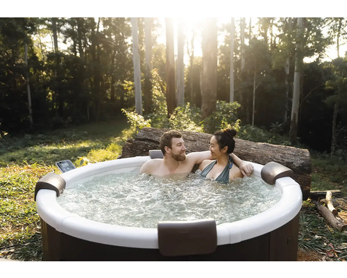Ein Paar entspannt in einem runden Außenwhirlpool im Garten mit Bäumen. Der Whirlpool verfügt über Kopfstützen und sprudelndes Wasser.