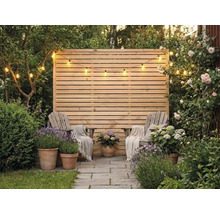 Sichtschutzzaun aus Holz mit horizontalen Lamellen, davor zwei Holzstühle mit Decken, Tontöpfe mit Lavendel und Hortensien sowie eine Lichterkette.