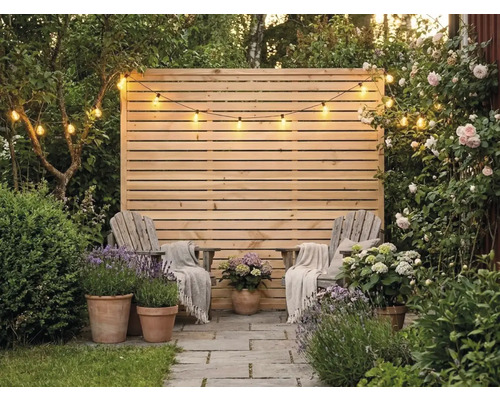 Sichtschutzzaun aus Holz mit horizontalen Lamellen, davor zwei Holzstühle mit Decken, Tontöpfe mit Lavendel und Hortensien sowie eine Lichterkette.