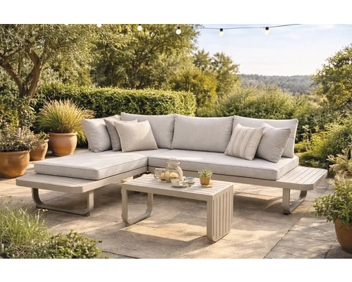 Modernes Garten-Lounge-Set aus Metall mit Stoffpolstern bestehend aus Ecksofa und Couchtisch auf einer gepflasterten Terrasse im grünen Garten.