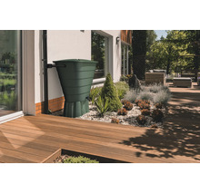Grüne Regentonne an einem Fallrohr neben einer Hauswand in einem modernen Garten mit Holzterrasse und Kiesbeet.
