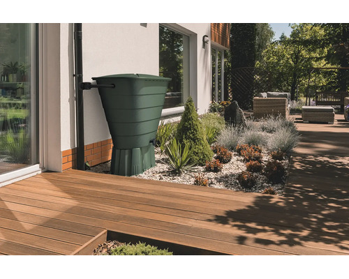 Grüne Regentonne an einem Fallrohr neben einer Hauswand in einem modernen Garten mit Holzterrasse und Kiesbeet.