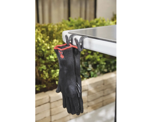 Schwarze Grillhandschuhe mit rotem Rand, mit Clips an einer Tischkante befestigt.