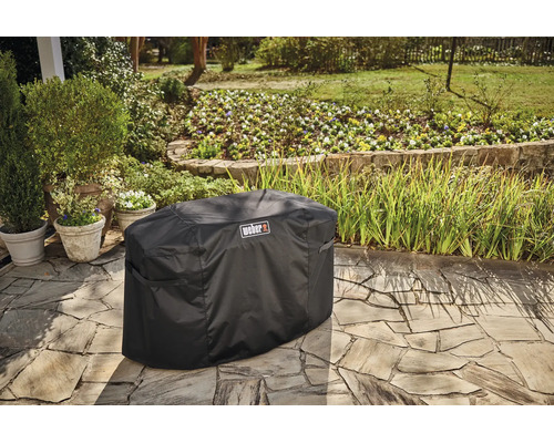 Schwarze Weber Grill-Abdeckhaube auf einer Steinterrasse in einem hellen, gepflegten Garten mit Topfpflanzen und Blumenbeeten.