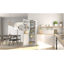 Moderne helle Küche mit offenem weißem Kühlschrank voller Lebensmittel, Arbeitsplatte aus hellem Holz und Essbereich im Hintergrund.