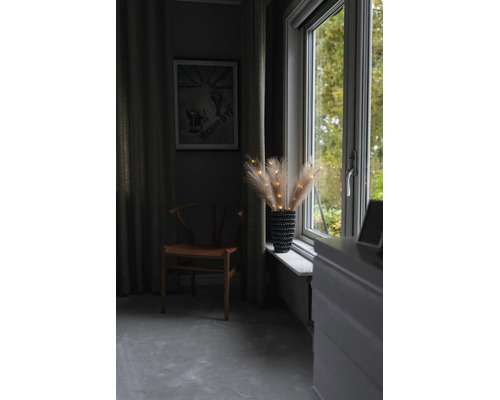 Beleuchtete Pampasgras-Zweige in einer Vase auf einem Fenstersims neben einem Holzstuhl in einem wohnlichen Zimmer.
