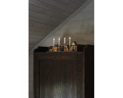 Weihnachts-Lichterbogen mit vier Kerzen und beleuchteter Dorfszene aus Holz auf einem dunklen Holzschrank vor einer holzgetäfelten Wand.